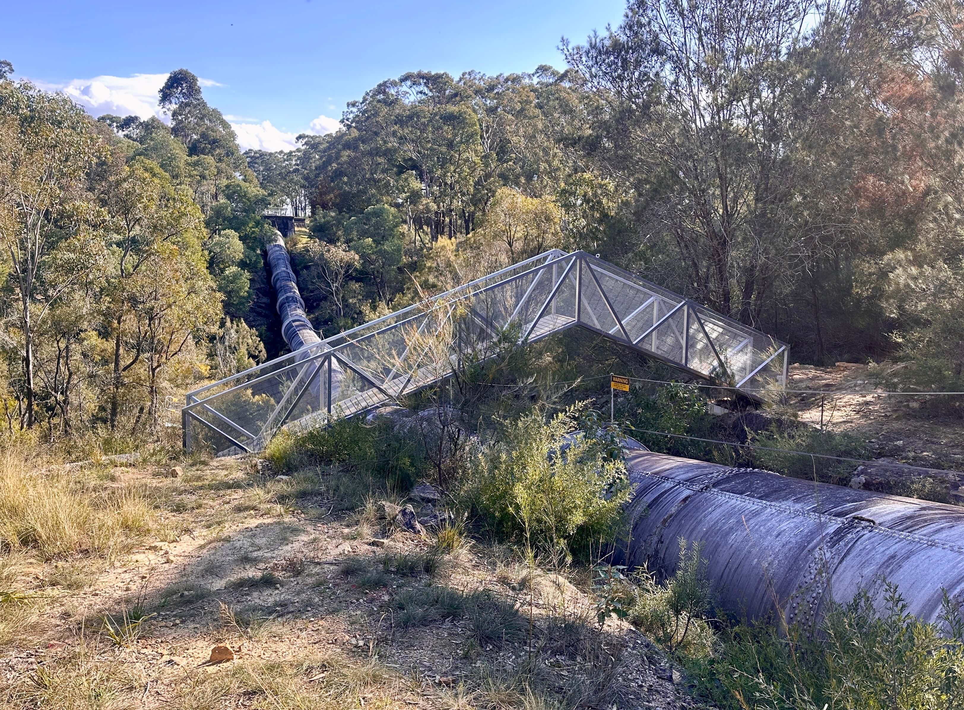 A metal and wooden bridge-like structure arches over a large metal pipeline, connecting bushland on either side of the pipe.